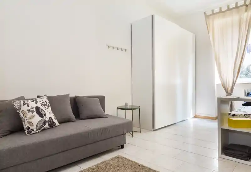Espace de vie lumineux et minimaliste avec un canapé gris, coussins décoratifs et une petite table d'appoint ; lumière naturelle et décoration neutre.