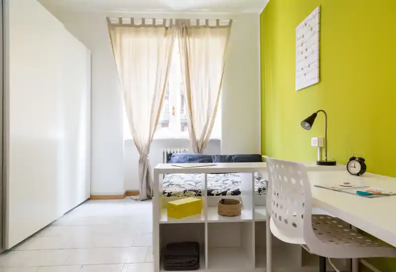 Bright, neatly staged bedroom with a single bed, desk and shelving; warm curtains and a green accent wall create a fresh, modern feel.