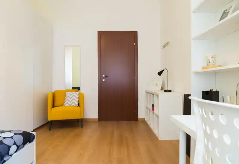 Bright, minimal bedroom corner with a warm wooden floor, a yellow accent armchair and a small visible portion of the bed; tidy shelving and a simple desk create a functional, modern feel.
