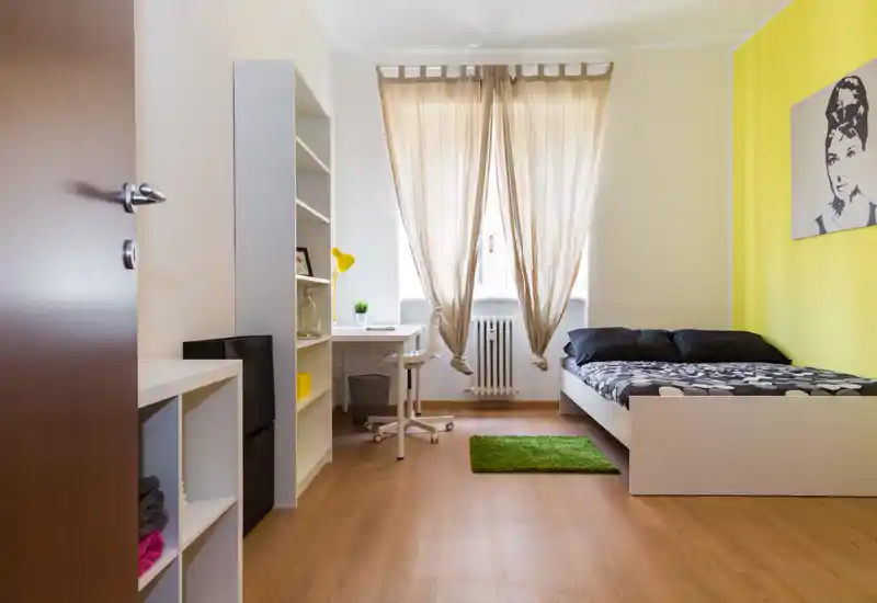 Bright, modern bedroom with a single bed, study desk and shelving; warm wooden floor and yellow accent wall create a welcoming, well-lit space ideal for a student or traveler.