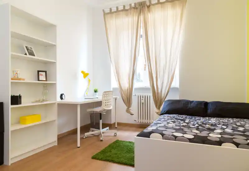 Bright, modern bedroom with a patterned double bed, desk and shelving — ideal for a student or professional. Natural light from the window and simple, cozy decor create a calm workspace and sleeping area.