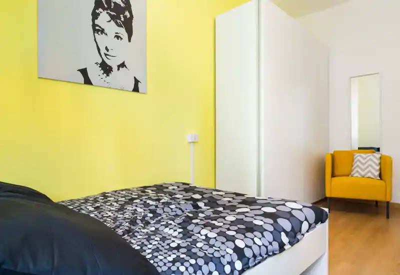 Bright, modern bedroom with a patterned duvet, yellow accent wall and a cozy mustard armchair; minimal white wardrobe provides storage and clean lines.