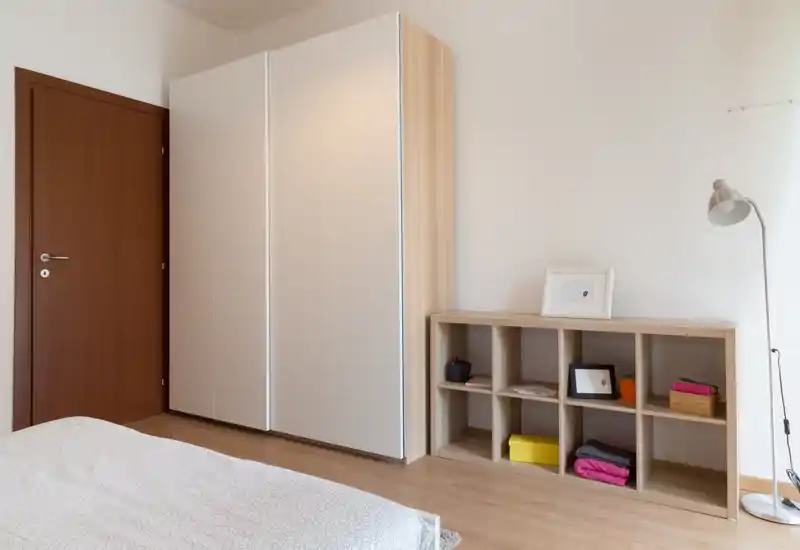Angolo di camera luminoso e minimale con un armadio scorrevole bianco, scaffale basso a cubi in legno e una lampada da terra; biancheria neutra e pavimento in legno creano un'atmosfera moderna e rilassante.