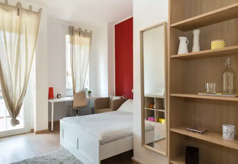 Camera da letto luminosa e moderna con letto bianco, luce naturale da ampie finestre, piccola scrivania e poltrona confortevole — perfetta per soggiorni comodi.