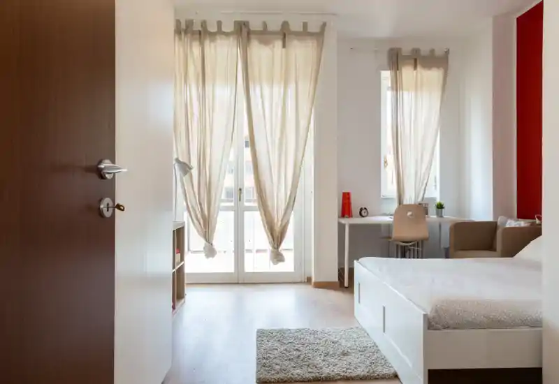 Camera da letto luminosa e ordinata con letto bianco, morbido tappeto e una scrivania vicino alla finestra; tende leggere diffondono la luce naturale creando un'atmosfera calma e accogliente.