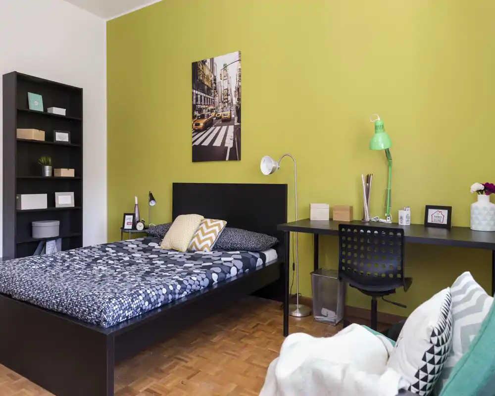 Camera da letto luminosa e moderna con letto matrimoniale, lampada da comodino, scrivania e scaffale. Parete verde come accento e tessili con pattern per un'atmosfera accogliente e contemporanea.