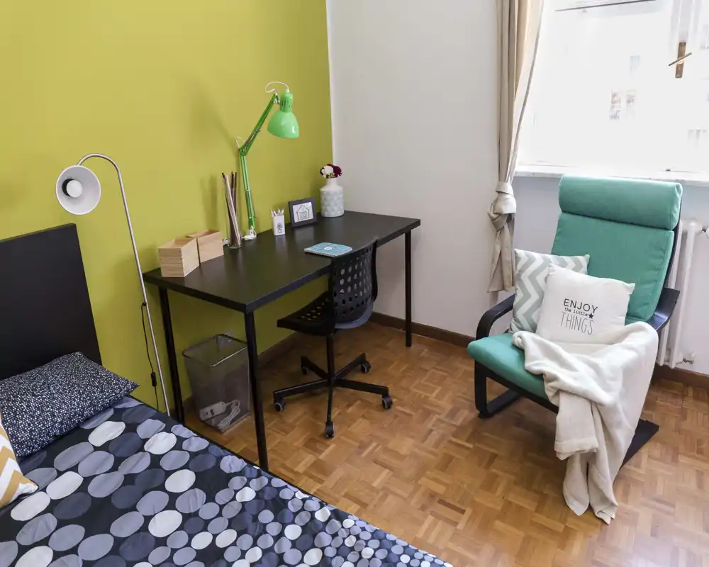 Camera da letto luminosa e accogliente con letto singolo e biancheria a fantasia, scrivania nera con lampada e sedia, e una poltrona turchese vicino alla finestra, perfetta per studiare e rilassarsi.