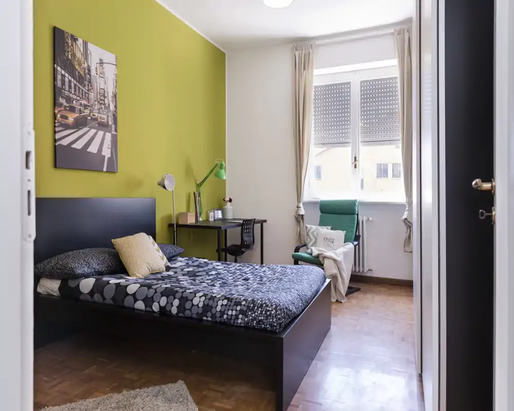 Camera da letto luminosa e di design con letto matrimoniale, lampade da comodino, scrivania e una poltrona comoda vicino alla finestra. Parete accentata verde e pavimento in parquet rendono l'ambiente accogliente.