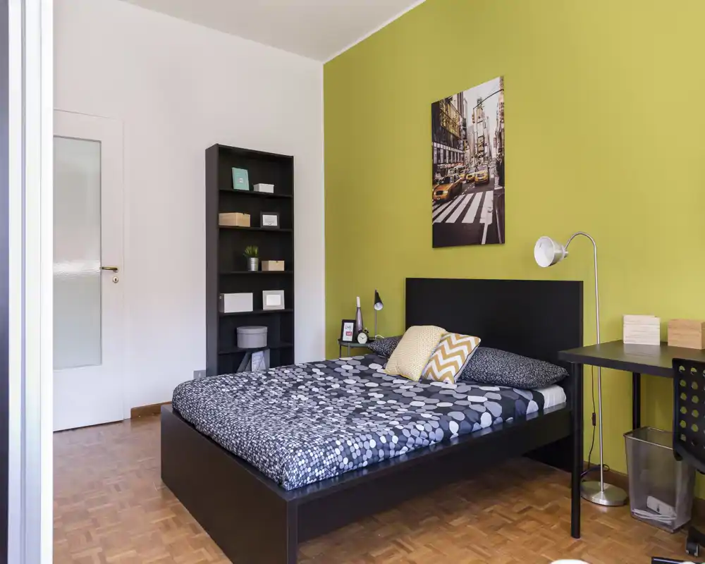 Camera da letto moderna e luminosa con letto matrimoniale, copriletto a fantasia e parete verde a contrasto. Comprende lampada da comodino, scaffale e scrivania — ambiente ordinato e ideale per annunci.