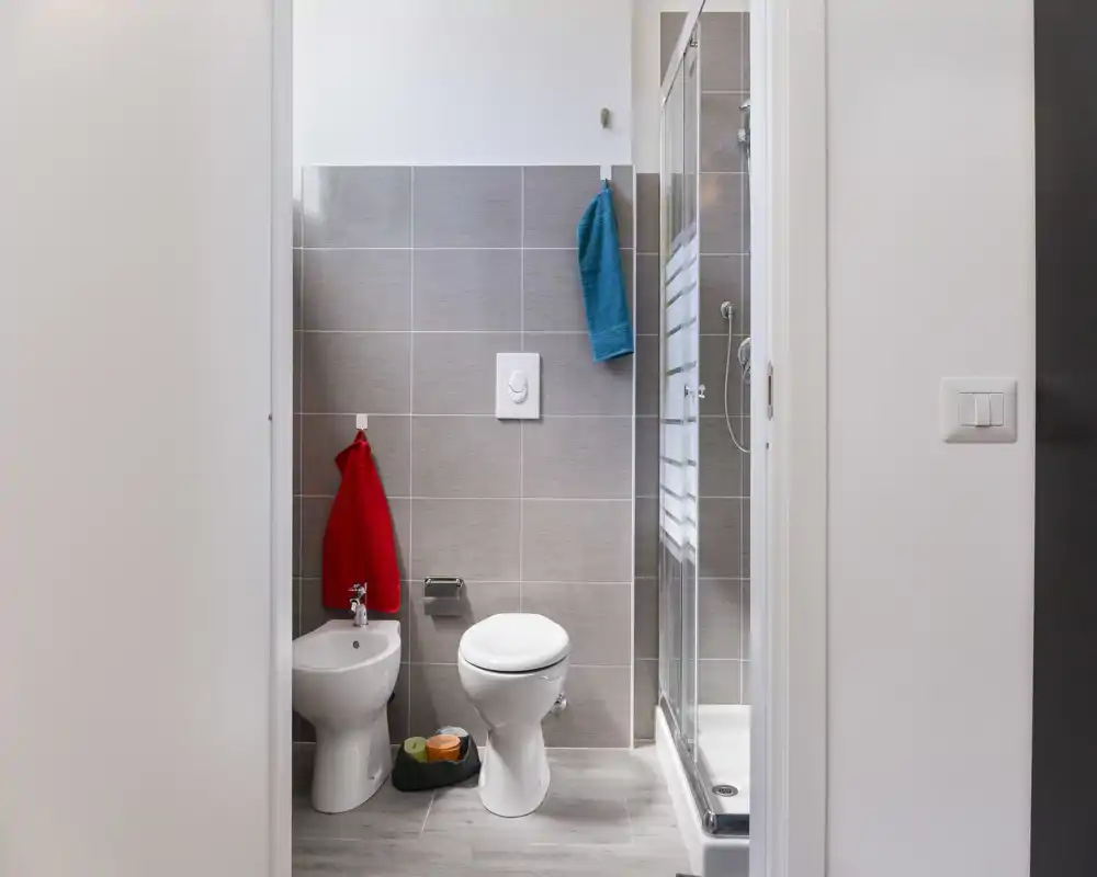 Bagno moderno compatto con wc, bidet e doccia con parete in vetro; piastrelle grigio chiaro e asciugamani colorati danno contrasto e un aspetto fresco.