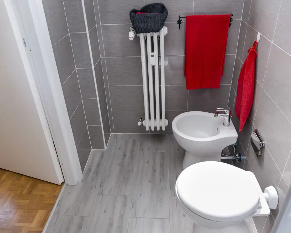 Piccolo bagno moderno con WC e bidet, pareti piastrellate grigie, pavimento effetto legno e asciugamani rossi che aggiungono un tocco di colore.