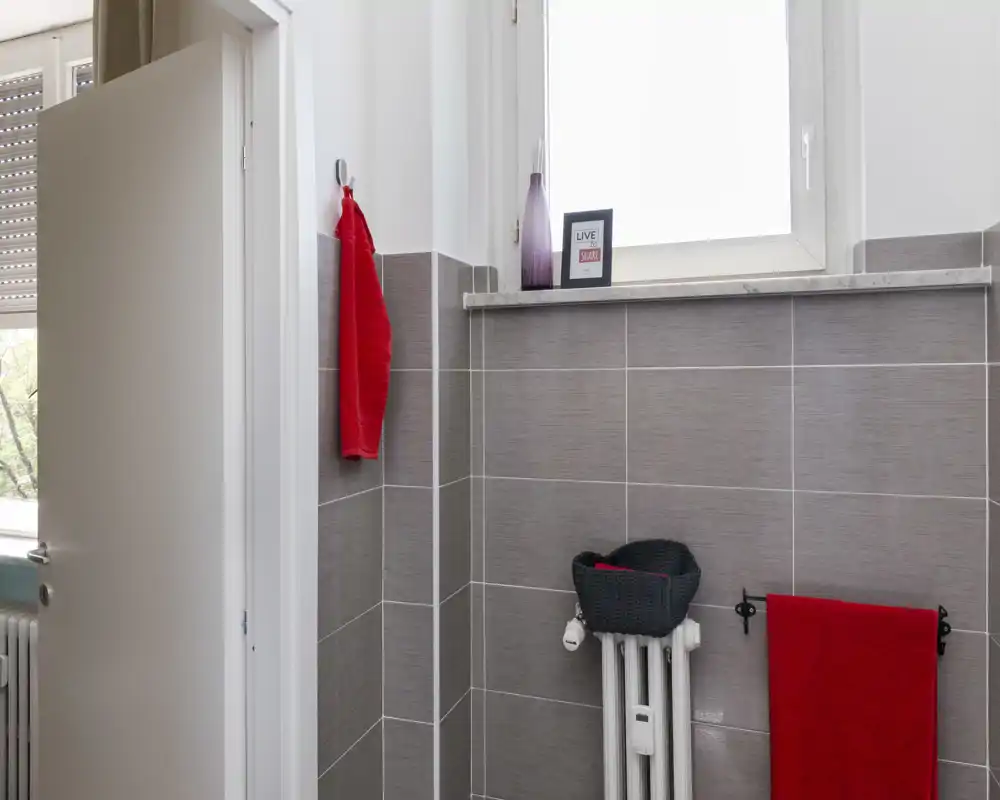 Angolo di bagno moderno con piastrelle grigie, termosifone con cesto in vimini e asciugamani rossi che danno un tocco di colore sotto una finestra satinata.