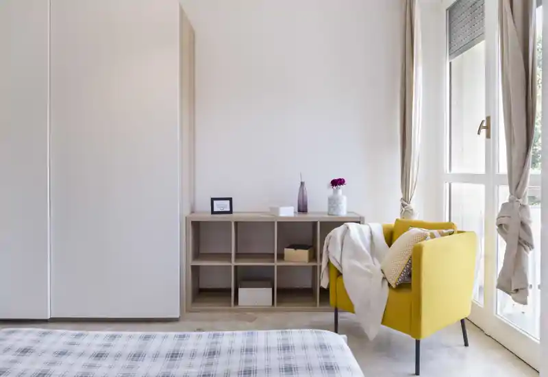 Angolo di camera luminoso e arredato con sobrietà: letto in primo piano, scaffale a cubi e una poltrona gialla vicino alla finestra che illumina l’ambiente.