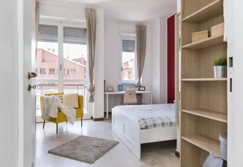 Dormitorio luminoso y bien decorado con una cama blanca visible, un escritorio junto a la ventana y un acogedor sillón amarillo junto a grandes ventanas que proporcionan mucha luz natural.