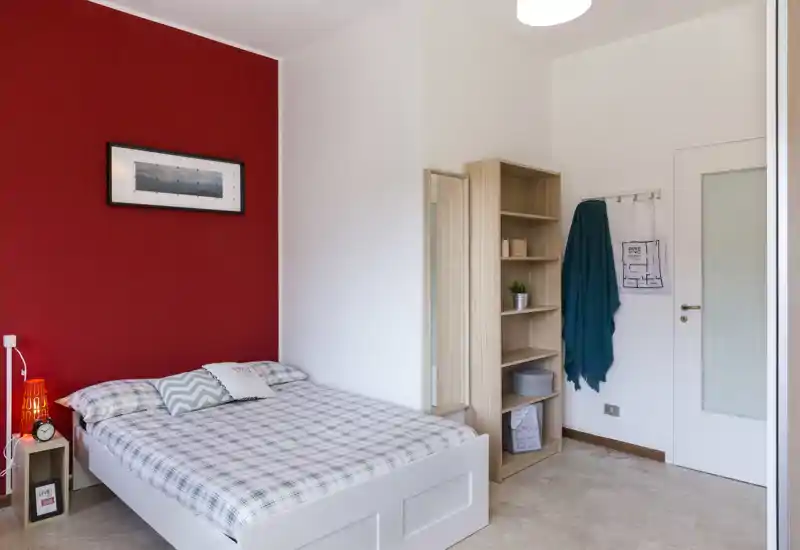 Camera da letto luminosa e ordinata con letto bianco davanti a una parete rossa a contrasto, comodino con lampada e mensole aperte per riporre oggetti — perfetta per foto annuncio che mettono in mostra comfort e arredo moderno.