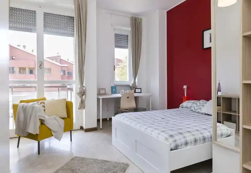 Camera da letto luminosa e arredata con letto bianco, biancheria a quadri e parete d'accento rossa; grandi finestre assicurano molta luce naturale e una poltrona gialla aggiunge un tocco di colore.