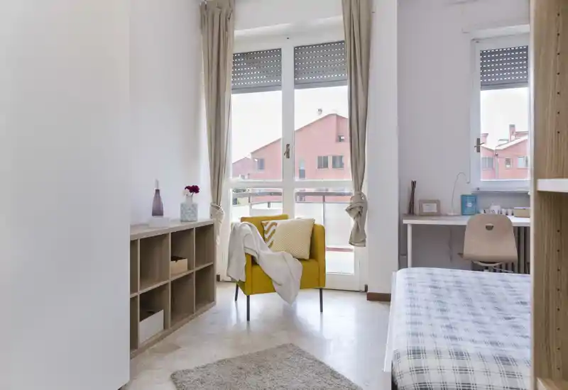 Camera da letto luminosa e ben allestita con il letto a quadretti visibile a destra, una poltrona gialla vicino alla finestra e una piccola scrivania — perfetta per annunci di affitto.
