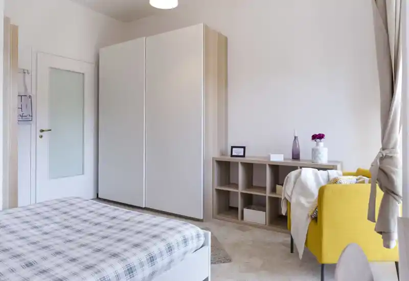 Camera da letto luminosa e moderna con letto ben rifatto, armadio bianco e una poltrona gialla vicino alla finestra — perfetta per un soggiorno accogliente.