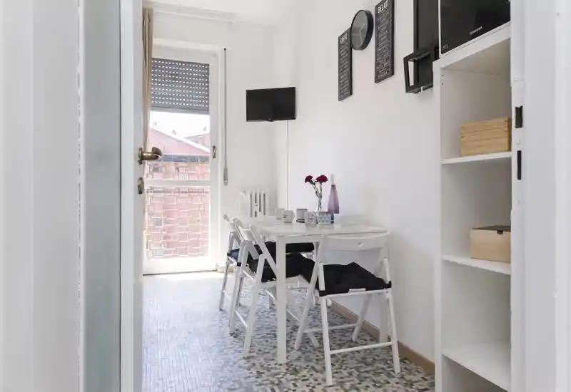 Área de comedor compacta y luminosa con mesa blanca y sillas plegables, decoración en la pared y luz natural desde la puerta al balcón — ideal para un apartamento pequeño.