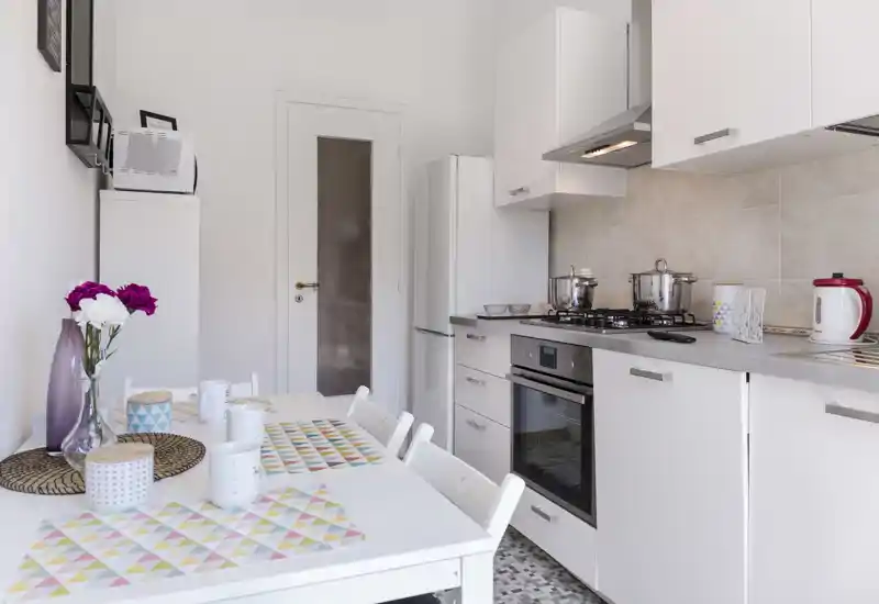 Cocina moderna y luminosa con armarios blancos, horno empotrado y placa de gas, pequeña mesa con manteles coloridos y un jarrón de flores.