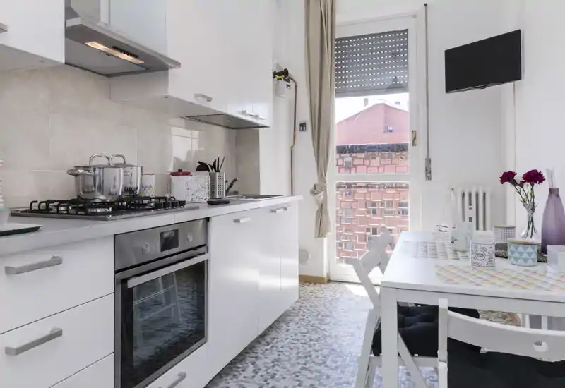 Cocina moderna y luminosa con muebles blancos, placa de gas y horno empotrado, además de una pequeña mesa junto a una ventana grande — ideal para apartamentos urbanos compactos.