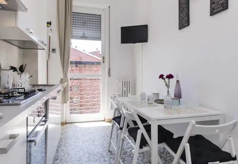 Cocina luminosa y moderna con electrodomésticos integrados y una pequeña mesa junto a la puerta del balcón; muebles blancos y detalles decorativos que crean un espacio limpio y acogedor.