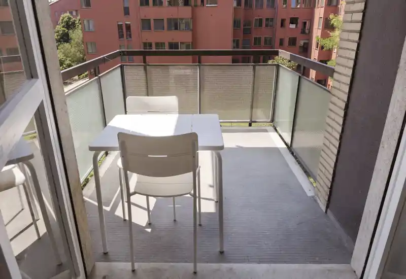 Balcón privado soleado con una pequeña mesa blanca y dos sillas, con vistas a los edificios vecinos. Ideal para desayunos o comidas ligeras al aire libre.