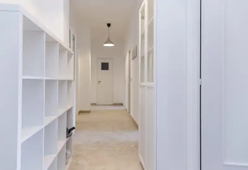 Entrada luminosa y minimalista con estanterías empotradas y lámpara colgante que conduce a puertas interiores; suelo de mármol y paredes blancas dan una sensación fresca y acogedora.