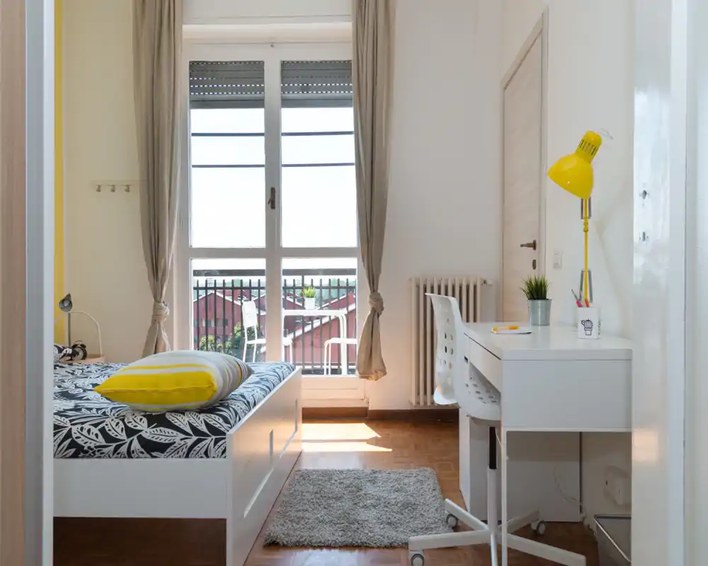 Camera da letto luminosa e stilosa con un comodo letto bianco, biancheria a fantasia e una finestra/terrazzino soleggiato; una scrivania bianca ordinata con una lampada gialla crea un angolo studio funzionale.