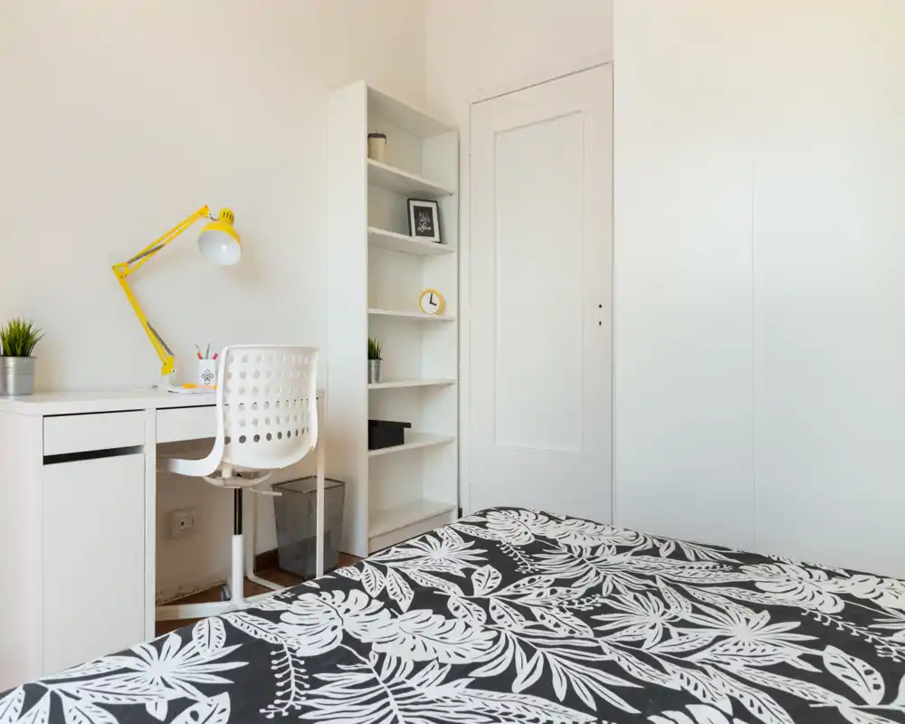 Camera da letto luminosa e minimal con copriletto a fantasia, scrivania bianca e lampada gialla — perfetta per una persona o uno studente.