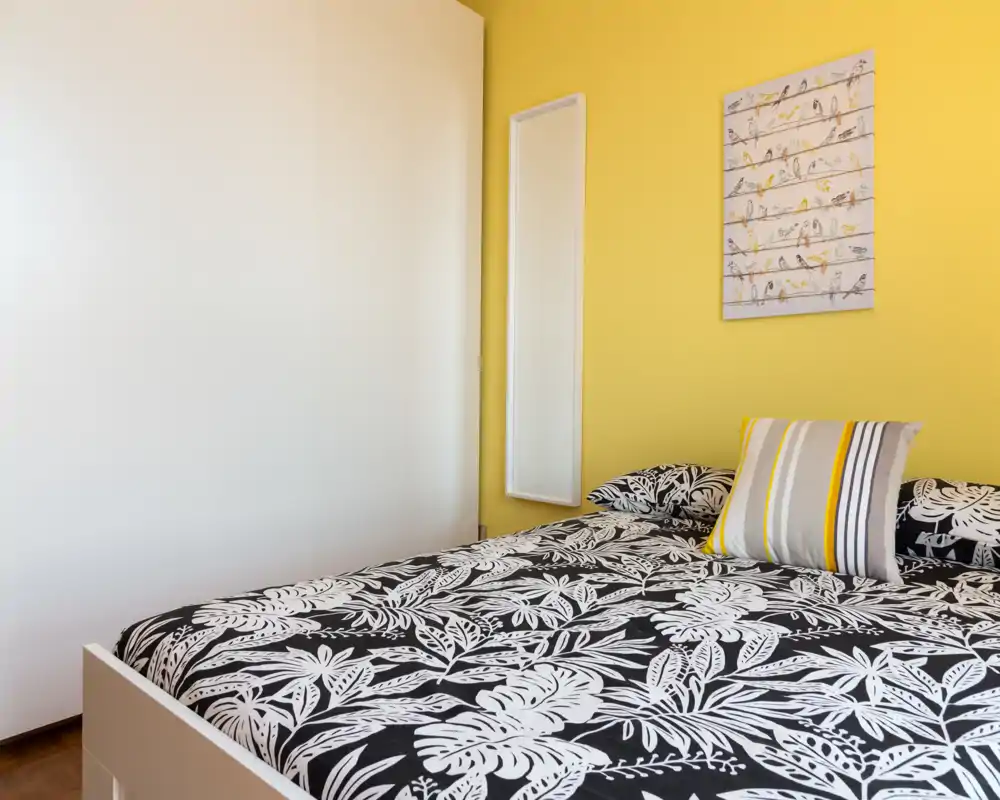 Camera da letto luminosa e moderna con copriletto nero e bianco a motivo, cuscino a righe come tocco d’accento e parete gialla calda. Linee pulite e arredo coordinato rendono la stanza fresca e accogliente.