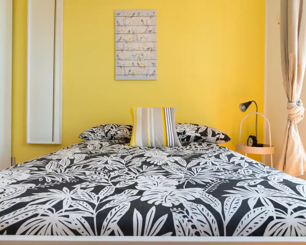 Camera da letto luminosa e di stile con parete gialla vivace, biancheria nera e bianca a fantasia e una lampada da comodino — ambiente accogliente e perfetto per foto di affitto.