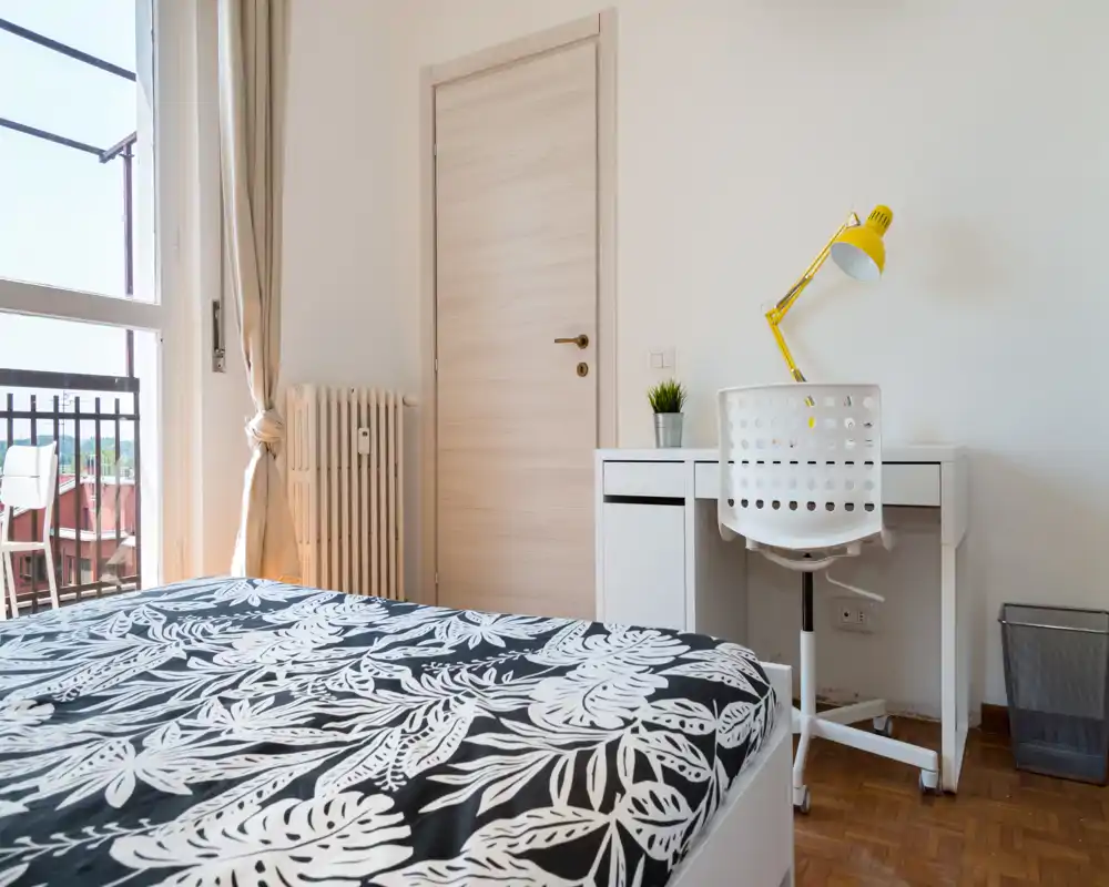 Camera da letto moderna e luminosa con copriletto a fantasia, scrivania con sedia e lampada gialla, e balcone che lascia entrare luce naturale.