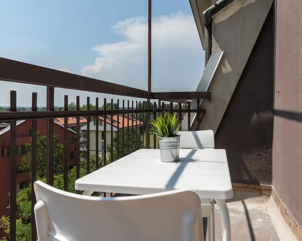 Balcone soleggiato e compatto con piccolo tavolo bianco e sedie, pianta in vaso sul tavolo e ringhiera in metallo con vista sugli edifici residenziali vicini.