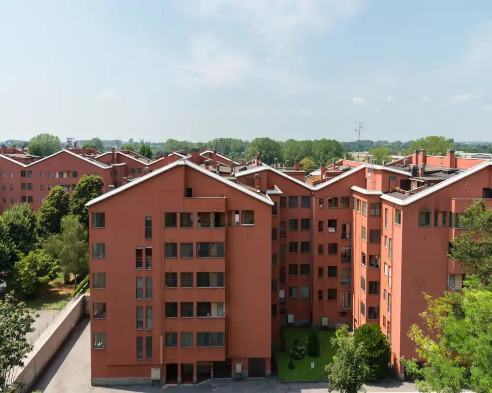 Vista esterna di un complesso di appartamenti rossi con più edifici, balconi e un cortile con vegetazione; scatto diurno chiaro utile per mostrare la facciata e l’intorno.
