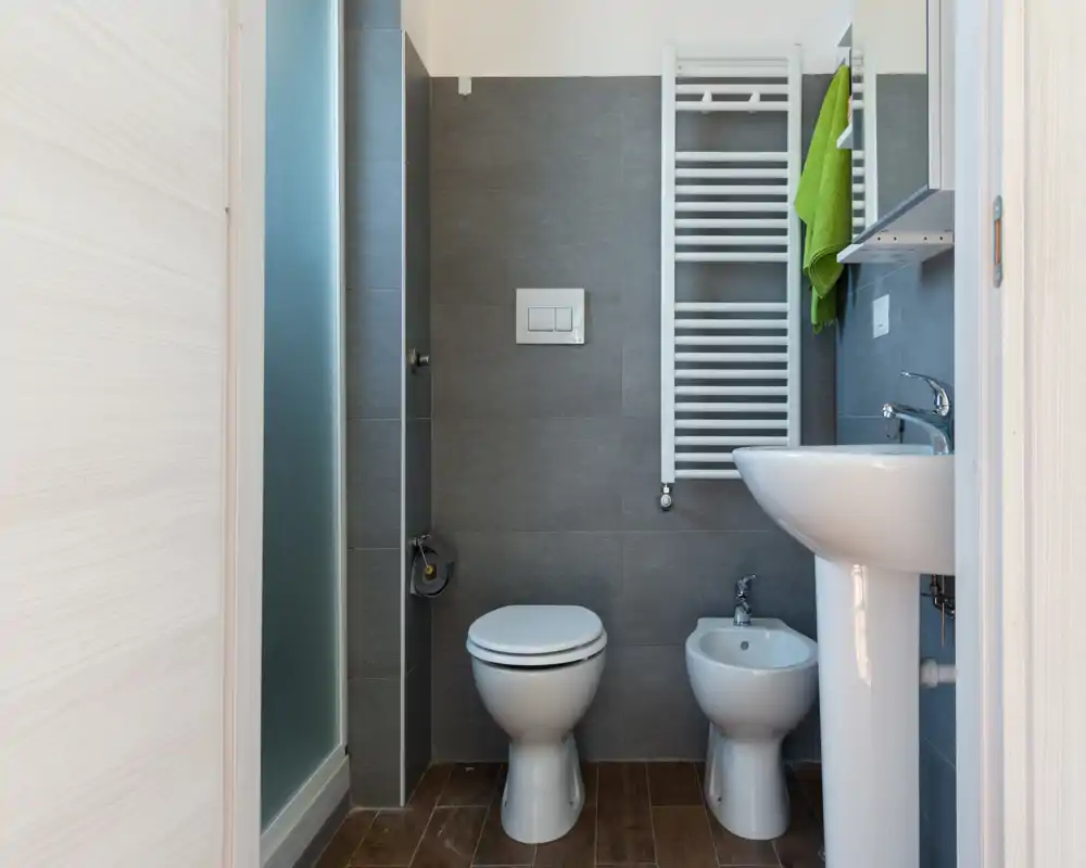 Bagno moderno compatto con placca di scarico a parete, WC, bidet e lavabo a colonna. Piastrelle grigie e pavimento effetto legno per un aspetto pulito e contemporaneo.