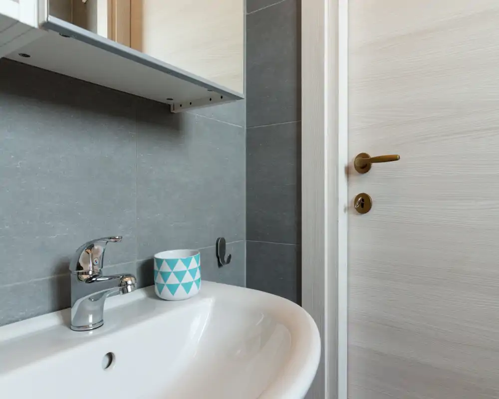 Piccolo lavabo bagno moderno con rubinetto cromato, rivestimento grigio e una tazza decorativa sul lavabo: pulito, minimal e ben illuminato.