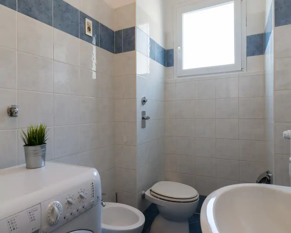 Bagno piastrellato compatto con WC, bidet e lavabo; una lavatrice è collocata sotto una piccola finestra che fornisce luce naturale. Piastrelle neutre con fascia decorativa blu, ambiente semplice e funzionale.