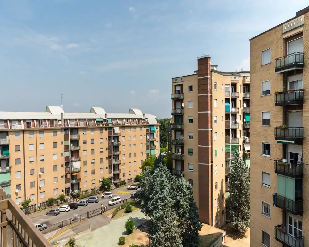 Vista urbana luminosa da balcone su condomini residenziali, parcheggi lungo la strada e alberi maturi: ideale per mostrare il quartiere e lo spazio esterno.