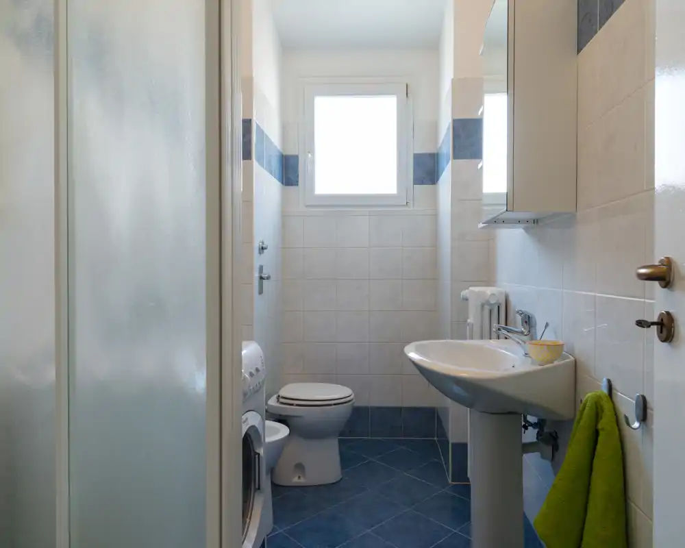 Bagno compatto e ben illuminato con piastrelle a parete e pavimento, lavabo su colonna, wc e box doccia a sinistra; pulito e funzionale per un affitto.
