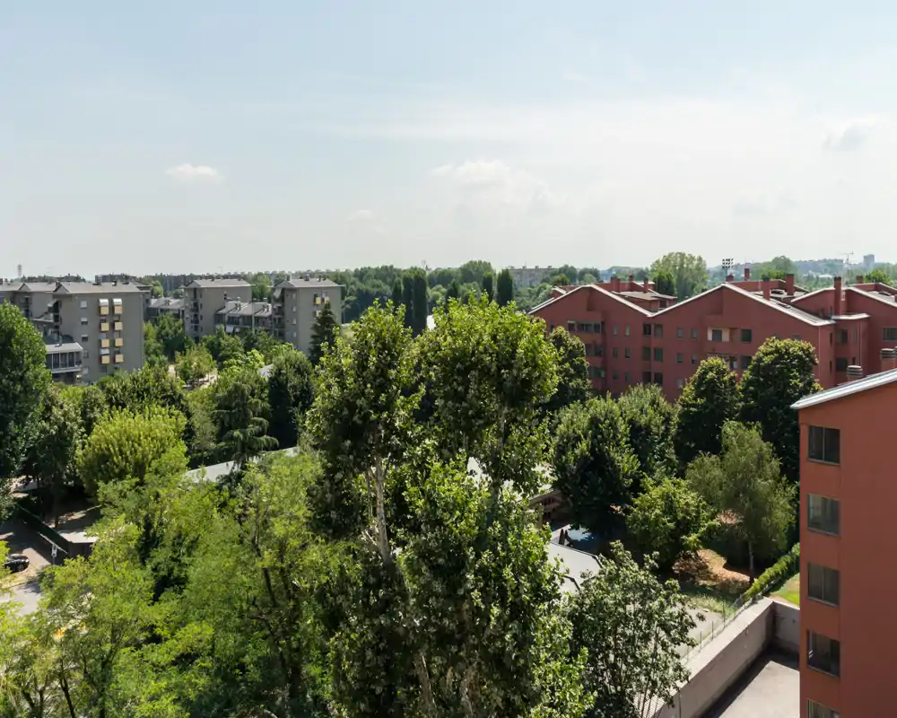 Vista luminosa da balcone/terrazzo su cortili alberati e condomini vicini, con cielo sereno e molta vegetazione.