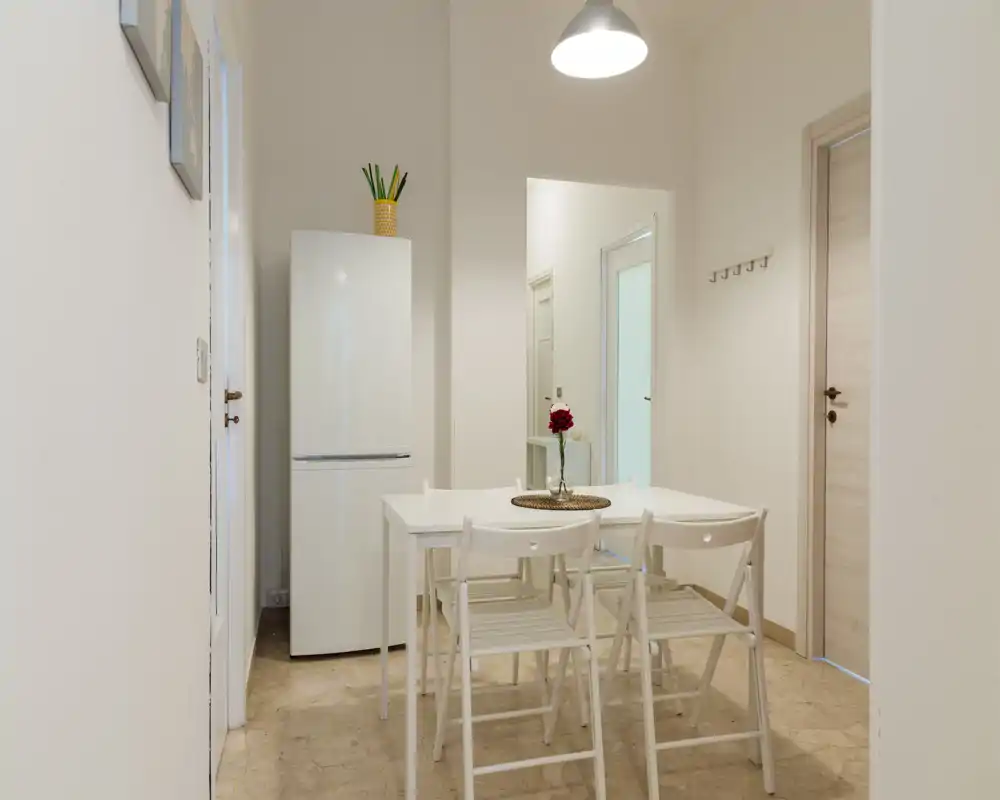 Zona pranzo compatta e luminosa con tavolo bianco e quattro sedie pieghevoli, valorizzata da un semplice centrotavola floreale: perfetta per pasti informali in un appartamento minimal.