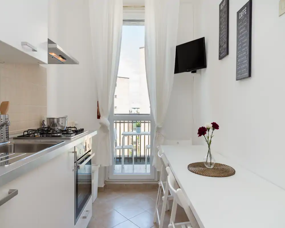 Cucina luminosa e compatta con piano cottura a gas, forno incassato e un lungo tavolo bianco vicino alla porta sul balcone. Le tende chiare e un piccolo vaso di fiori rendono l’ambiente fresco e accogliente.