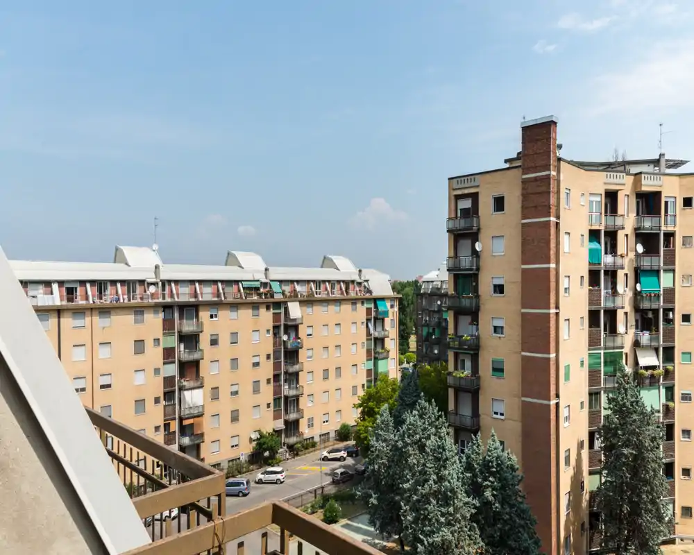 Vista dal balcone su condomini e una strada fiancheggiata da alberi; scatto diurno luminoso con cielo limpido e dettagli urbani.
