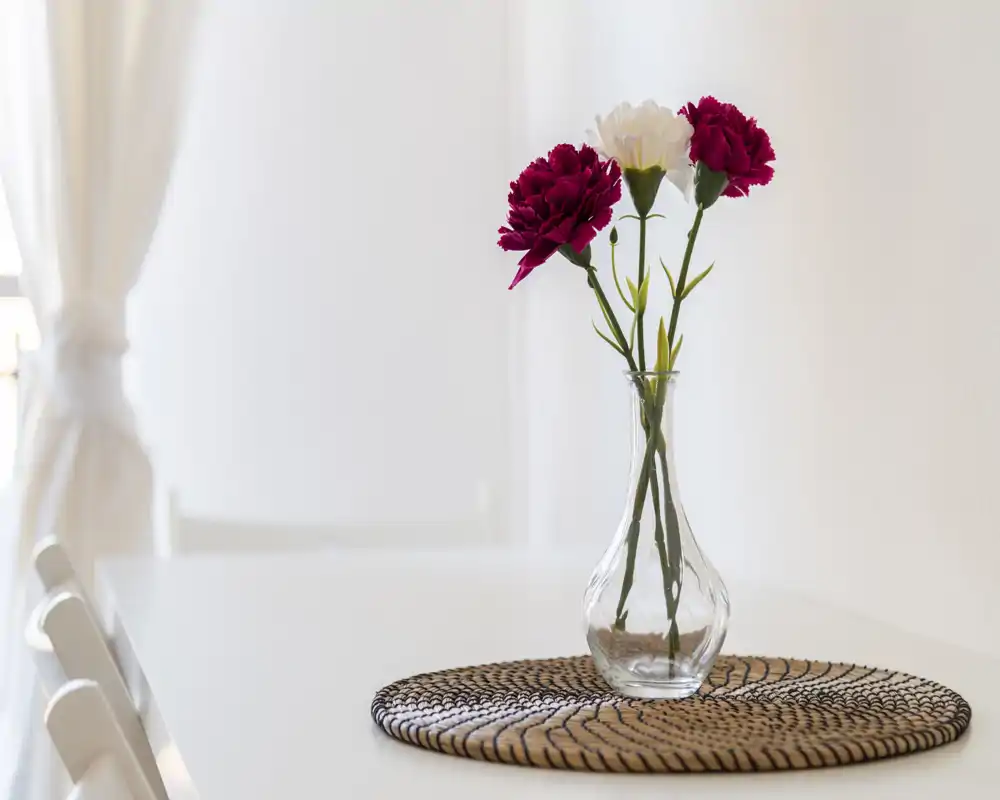 Scatto luminoso e minimal della zona pranzo con un vaso di fiori su un tavolo bianco e sedie nella luce naturale soffusa.