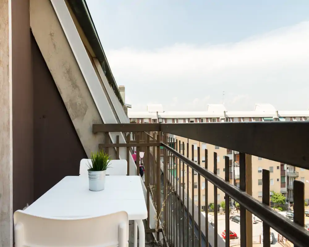 Balcone soleggiato e stretto con piccolo tavolo bianco, due sedie e una piantina in vaso, con vista sugli edifici vicini — perfetto per il caffè mattutino.
