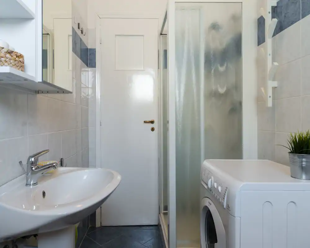Bagno compatto e pulito con lavabo, doccia con box e lavatrice — pareti piastrellate chiare e rubinetteria moderna creano uno spazio funzionale e curato.