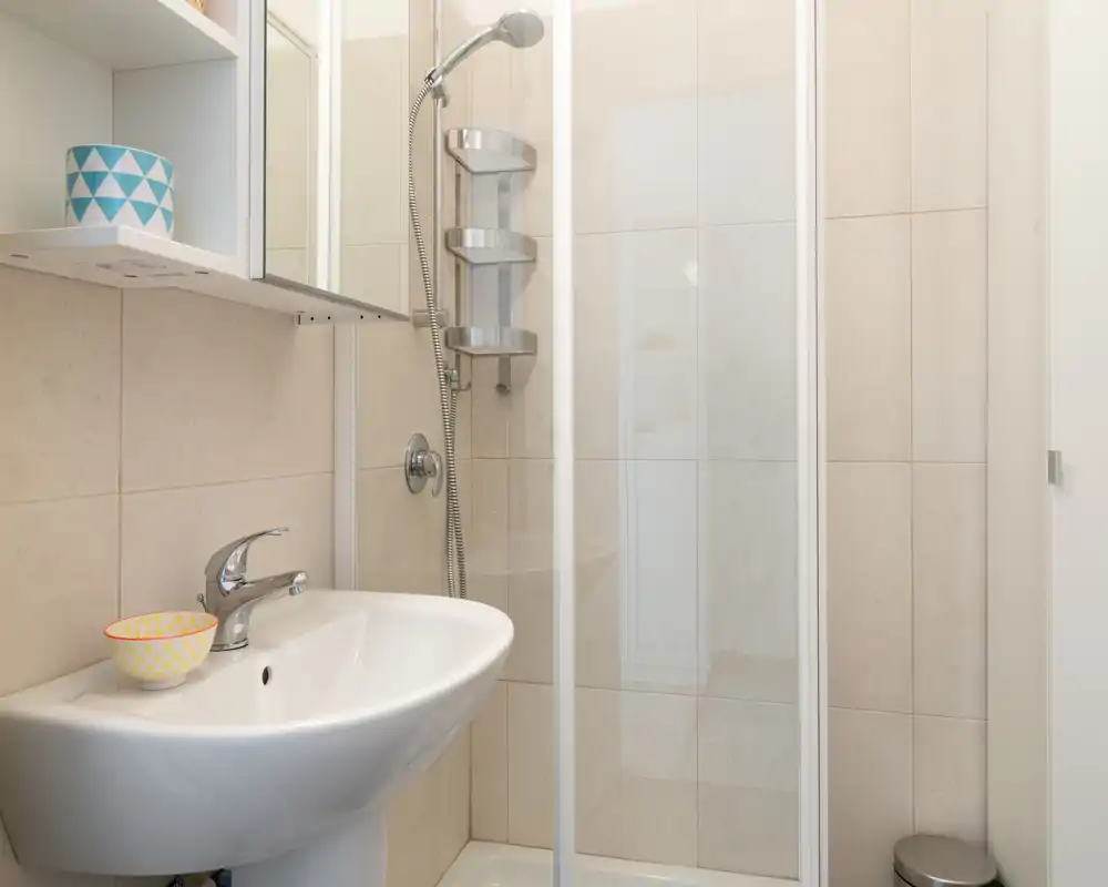 Bagno luminoso e pulito con lavabo a colonna e box doccia in vetro dotato di doccetta e mensoline da parete.