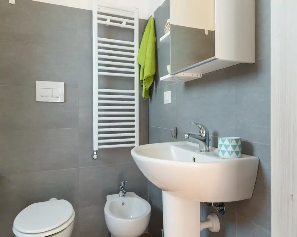 Bagno moderno compatto con lavabo a colonna, bidet e wc, piastrelle grigie neutre e termoarredo. Luminoso e pulito, perfetto per un annuncio di affitto.