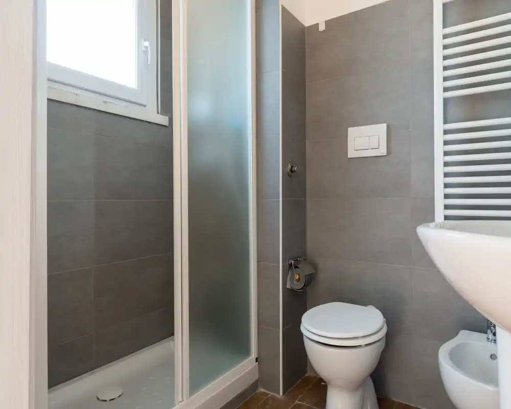 Bagno moderno e compatto con cabina doccia, WC e lavabo a parete; piastrelle grigie neutre e termoarredo a scala per un aspetto pulito e minimalista.